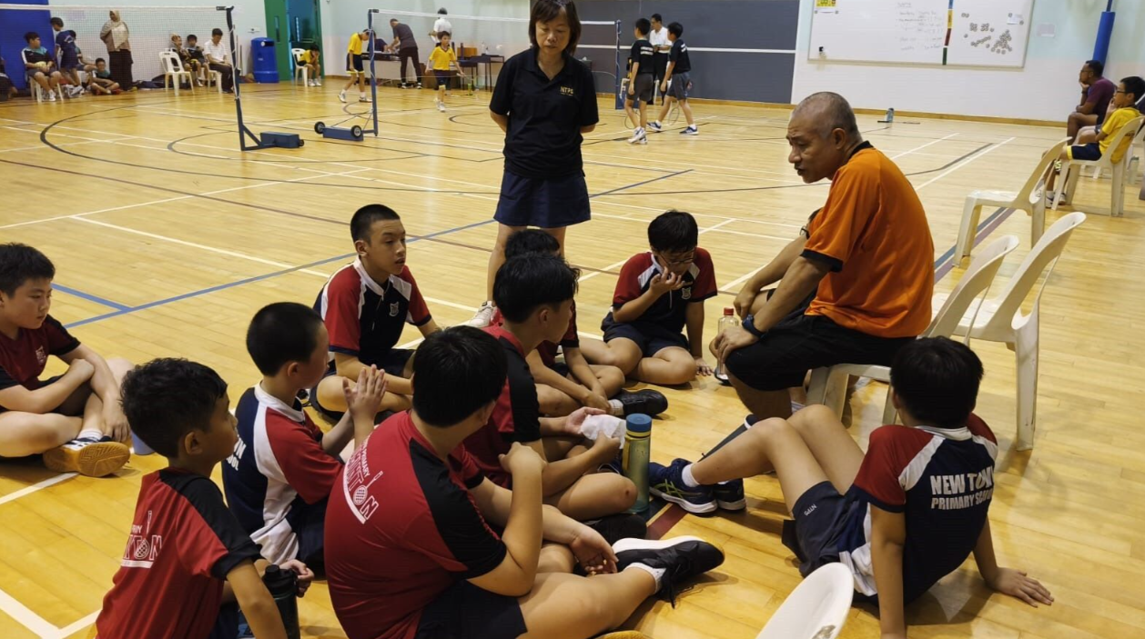 Two coaches talk to a group of young students sitting on a sports hall floor during a badminton session.