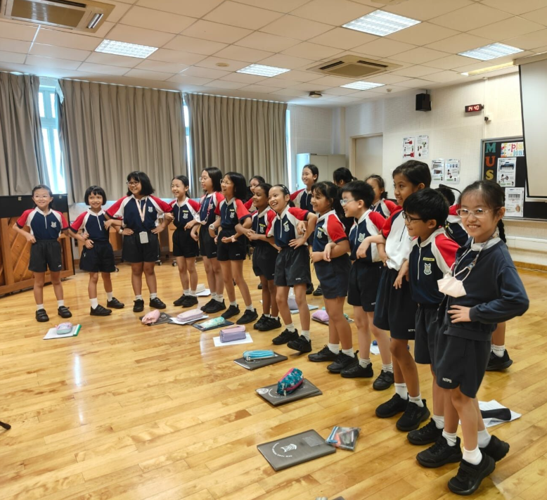 A group of young students in navy and red uniforms stand smiling in a row on a wooden floor indoors.