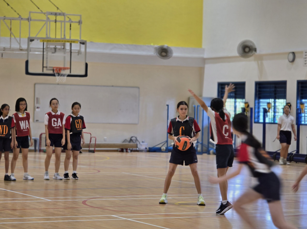 Girls playing netball indoors, one holding an orange ball while teammates in red and navy bibs stand nearby.
