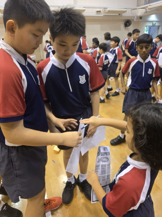 Three children in matching school uniforms examine a paper sheet in a gym with more students in the background.