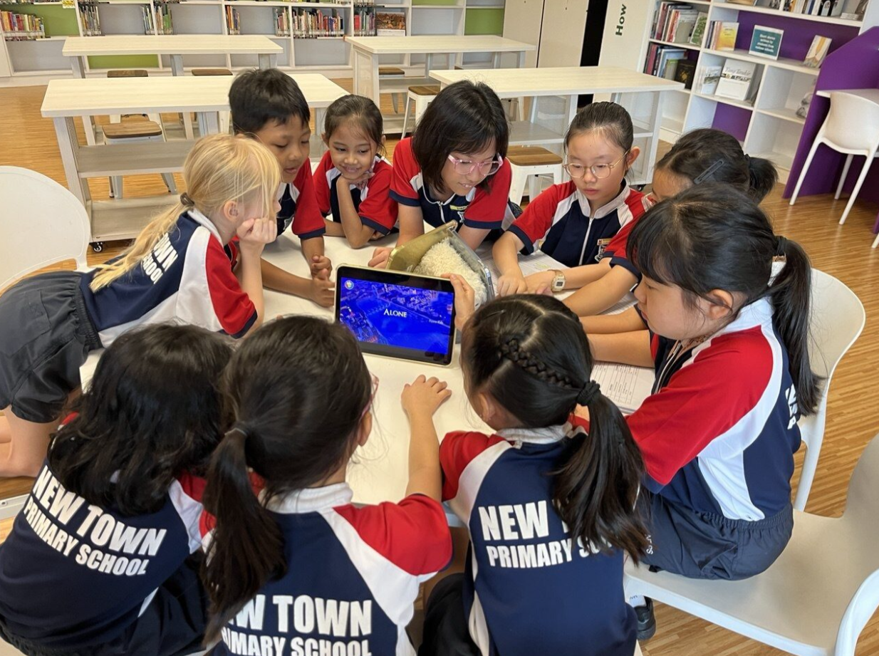 New Town Primary School students in uniform huddle around a tablet displaying a blue screen in a library.