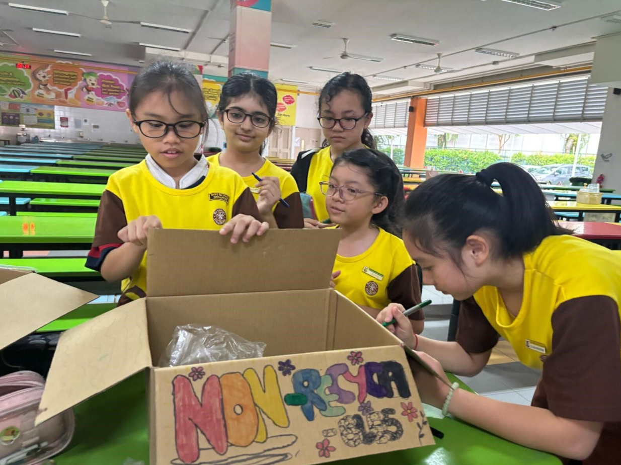 Five girls in yellow school uniforms gather around a decorated cardboard box with colorful lettering in a school cafeteria.