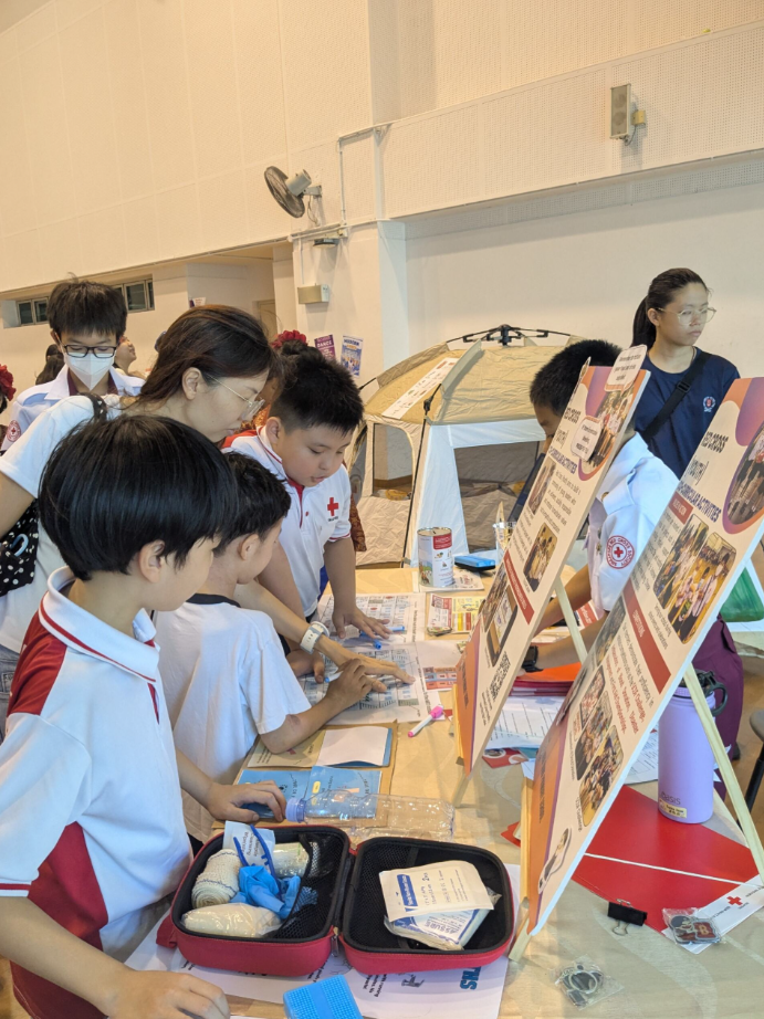Students in Red Cross uniforms gather around a table with first aid kits and informational boards at an indoor event.