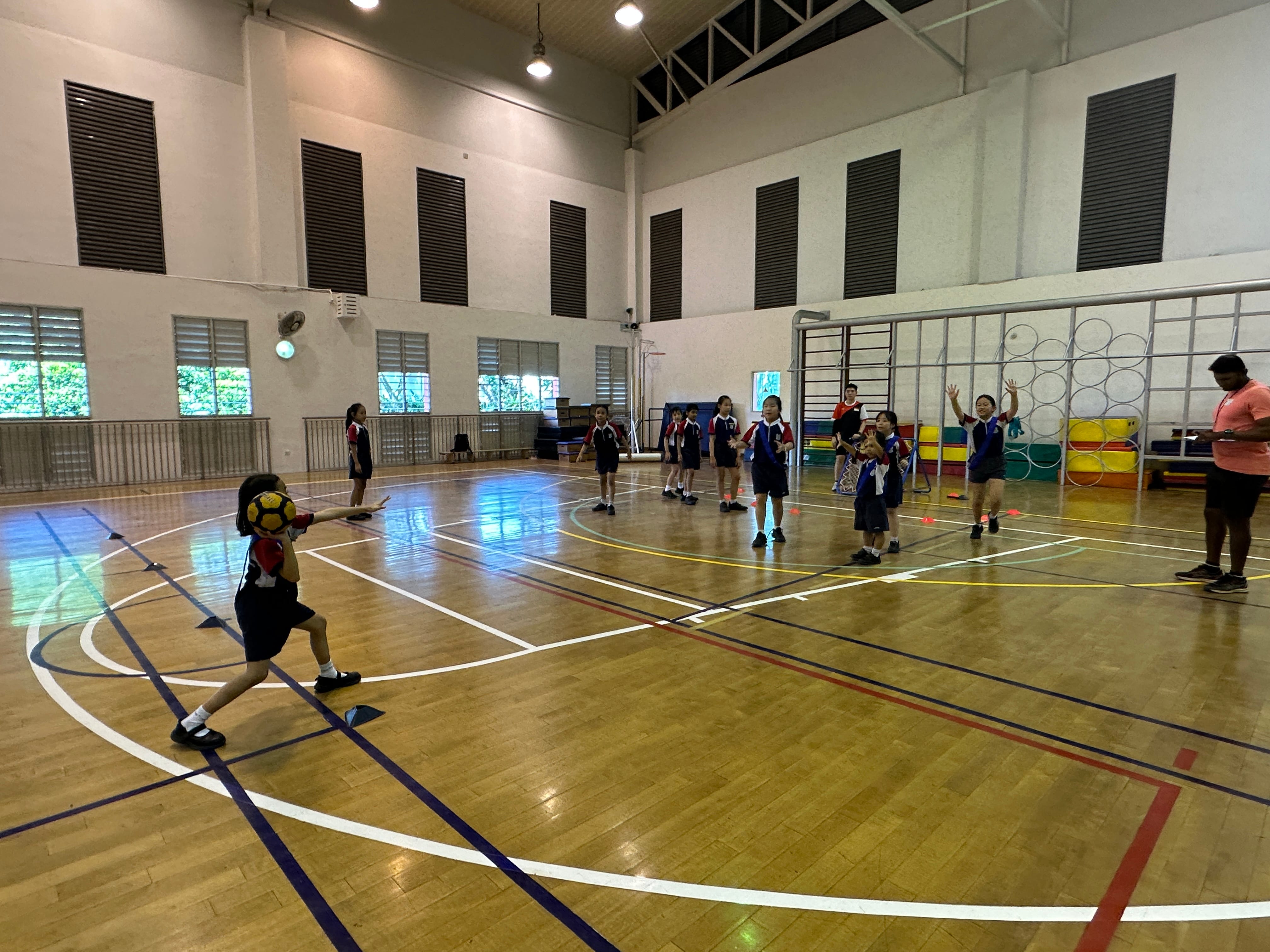 Children in uniforms play with a ball in an indoor gymnasium with wooden floors and a teacher watching nearby.