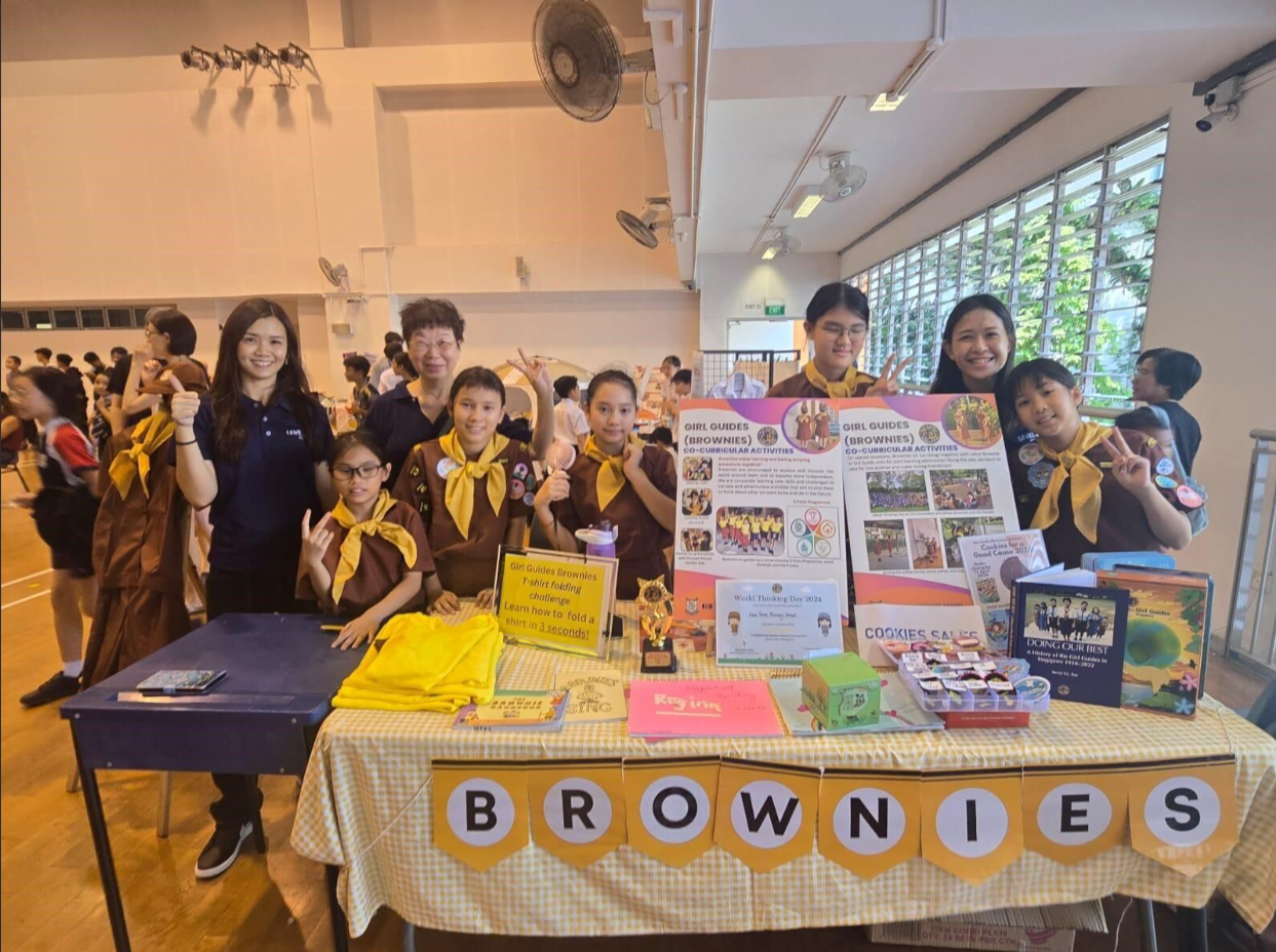 Girl Guides Brownies members in brown uniforms pose at a display table with posters and cookies in a hall.