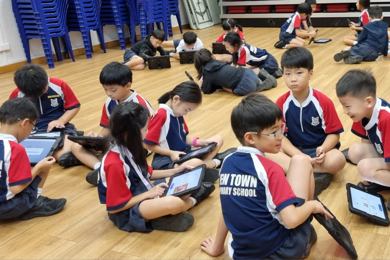 Primary school children in uniforms sit on a wooden floor using tablets during a group activity.