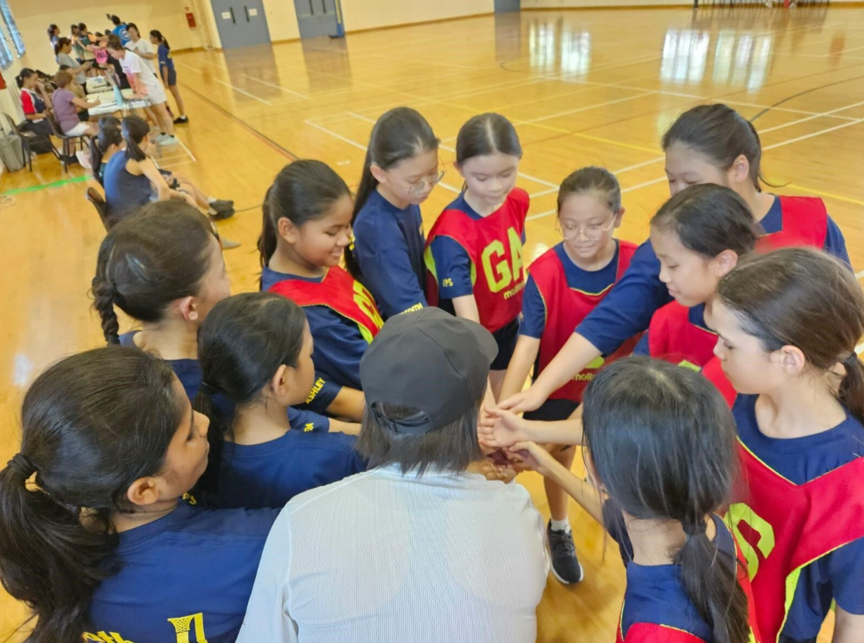 A group of young girls in blue and red sports jerseys form a team huddle with hands stacked together in a gymnasium.