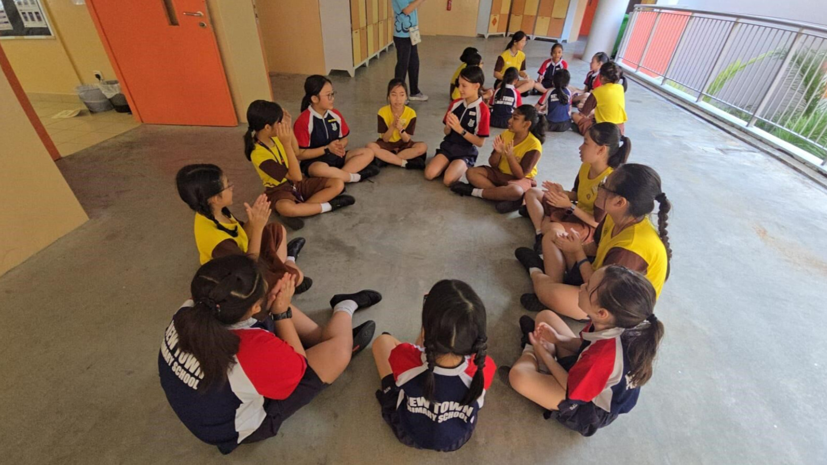 Students in school uniforms sit in a circle on a corridor floor, clapping hands in a group activity.