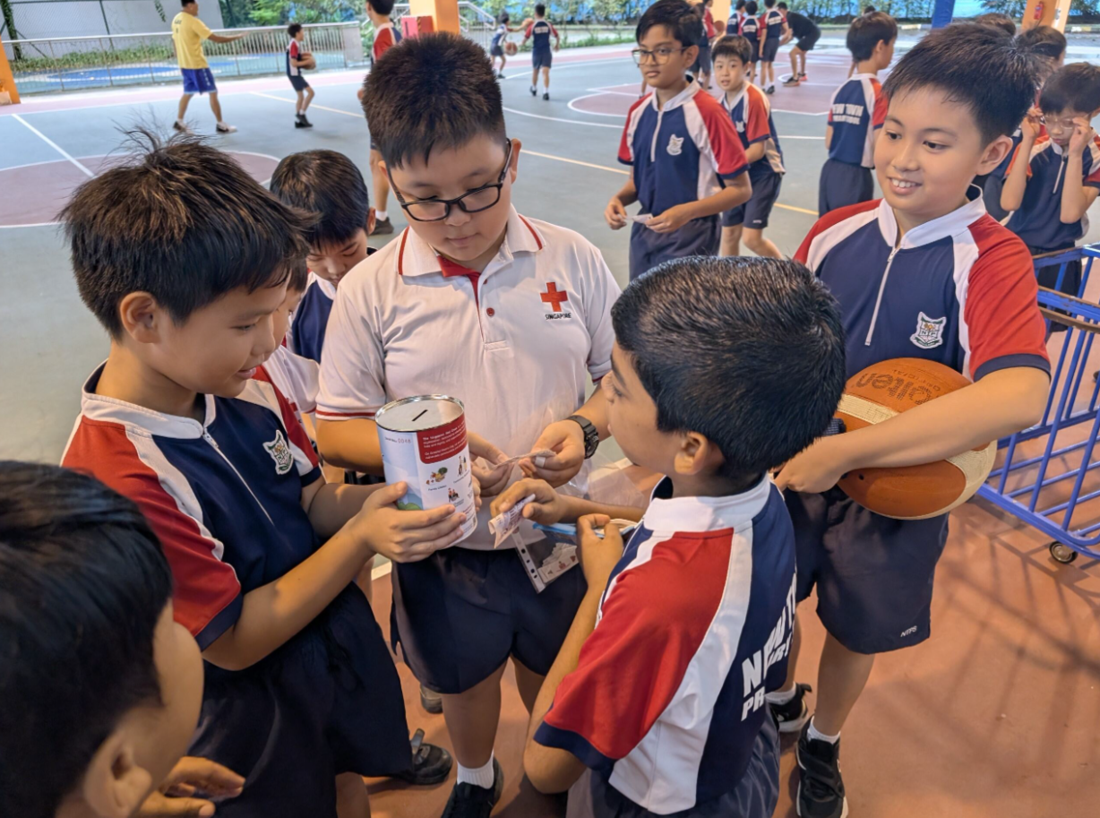 A boy in a Red Cross shirt collects donations in a tin from uniformed schoolboys on an outdoor court.