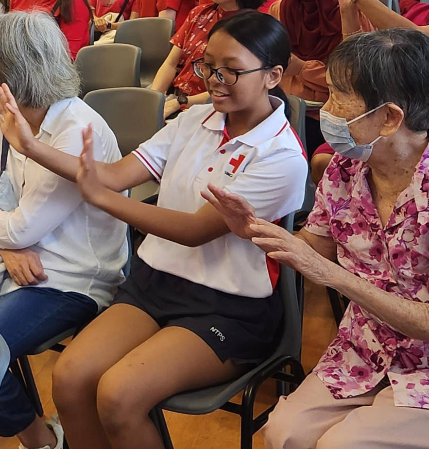 A young girl in a Red Cross uniform claps hands with an elderly masked woman while seated among others.