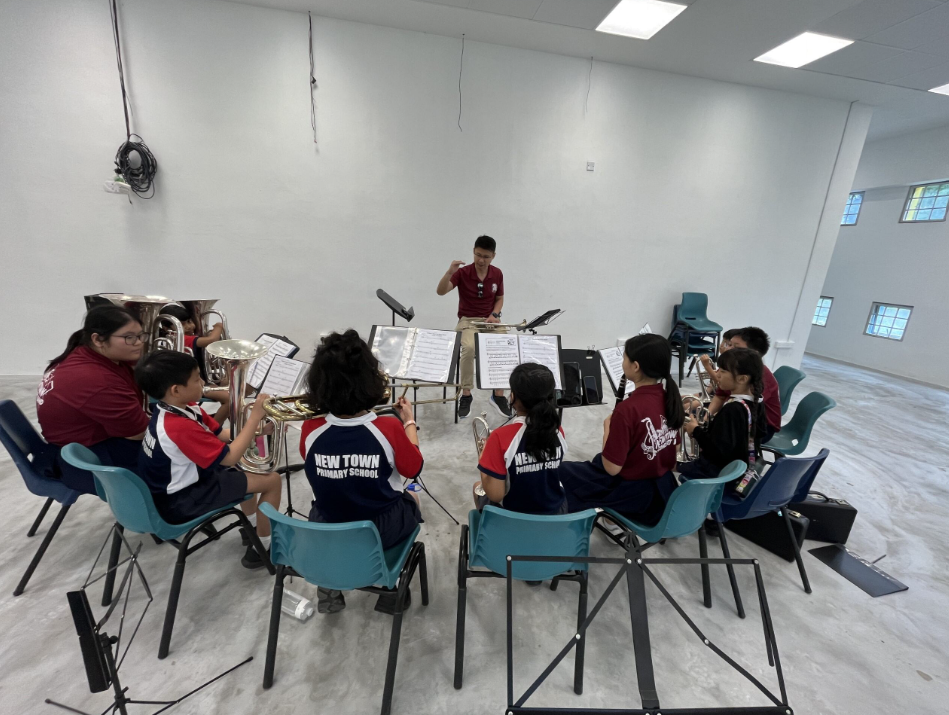 A conductor leads New Town Primary School students playing brass instruments in a circle in a rehearsal room.