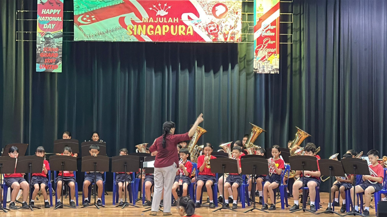A conductor leads a children's brass band on stage beneath Majulah Singapura National Day banners.