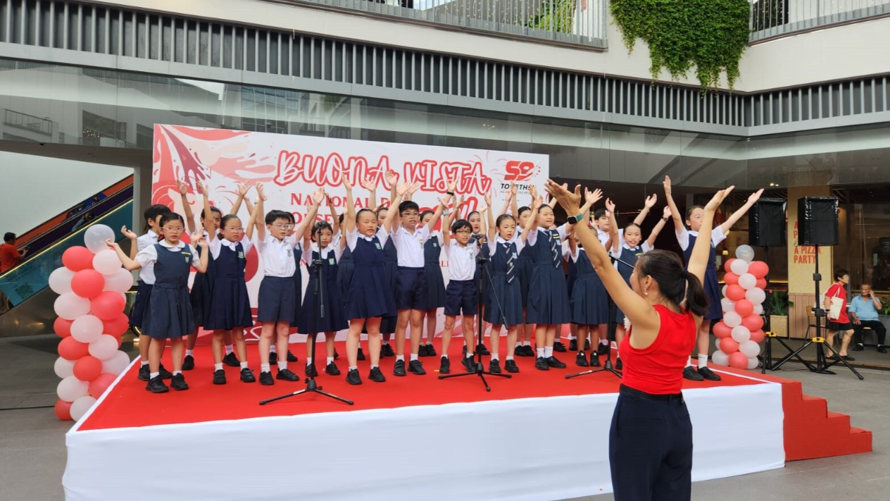 Children in school uniforms perform on a red stage at Buona Vista National Day event, led by a conductor in red.