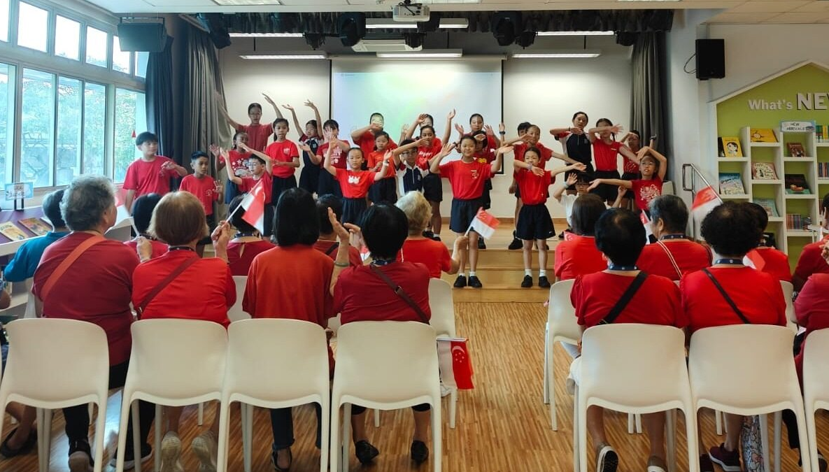Children in red shirts perform on a stage with raised arms before a seated audience holding small Singapore flags.