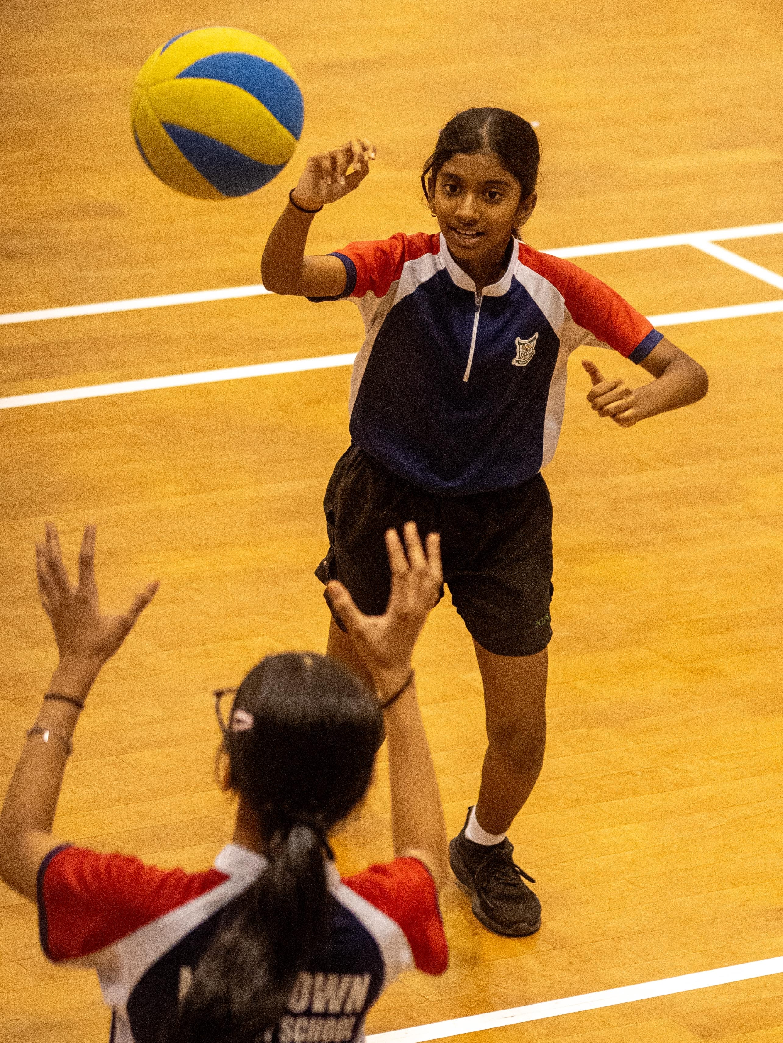 A girl in a blue and red uniform hits a yellow and blue volleyball toward a teammate on a wooden indoor court.