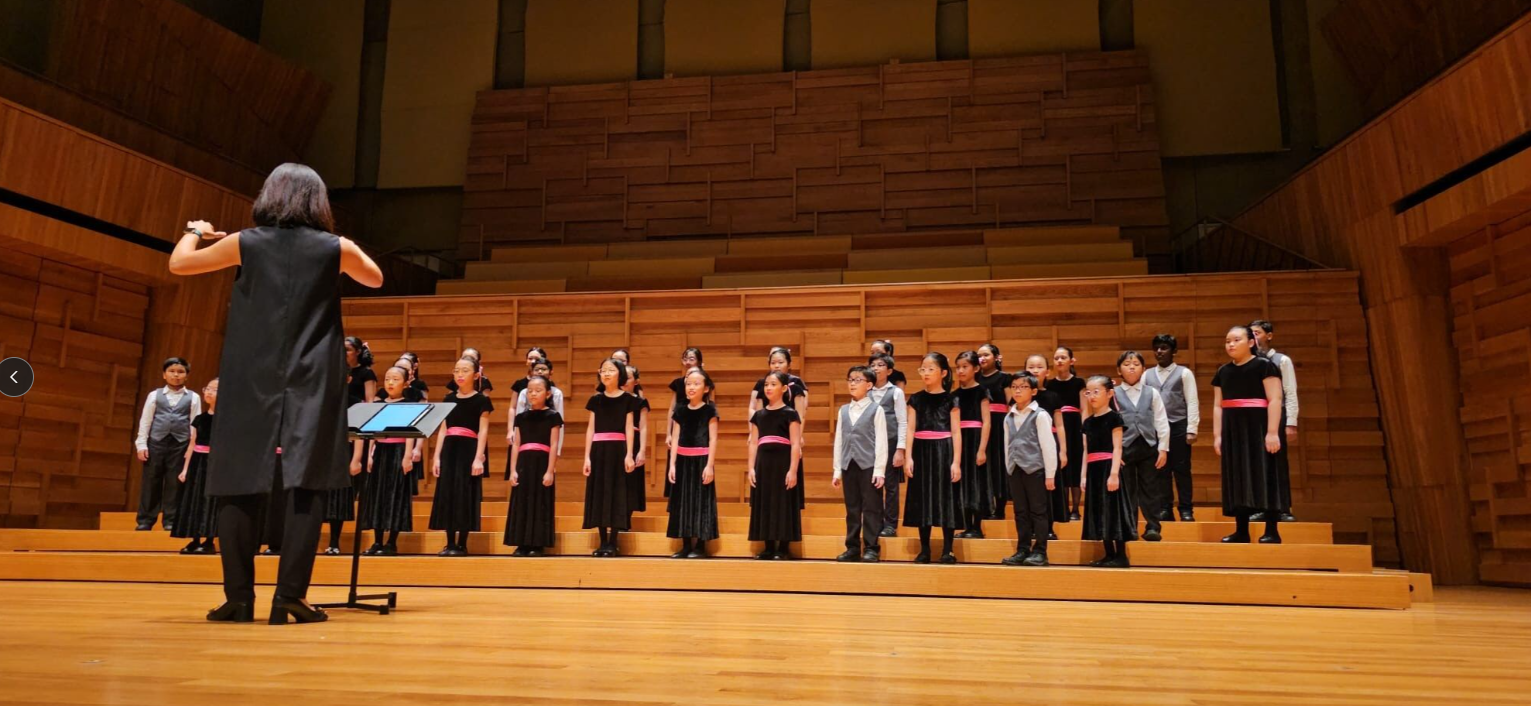 A conductor in black leads a children's choir in black uniforms with pink sashes on a wooden concert hall stage.