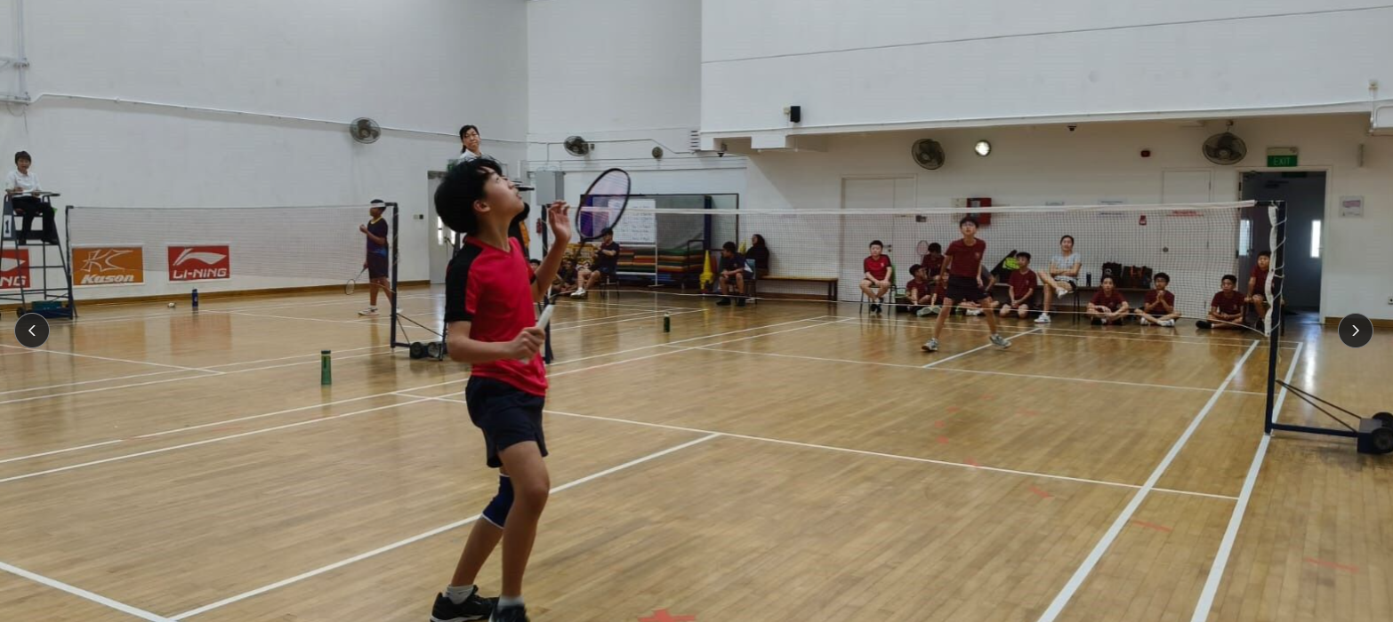 A young boy in red holds a badminton racket looking up on an indoor court, with students watching from the sideline.
