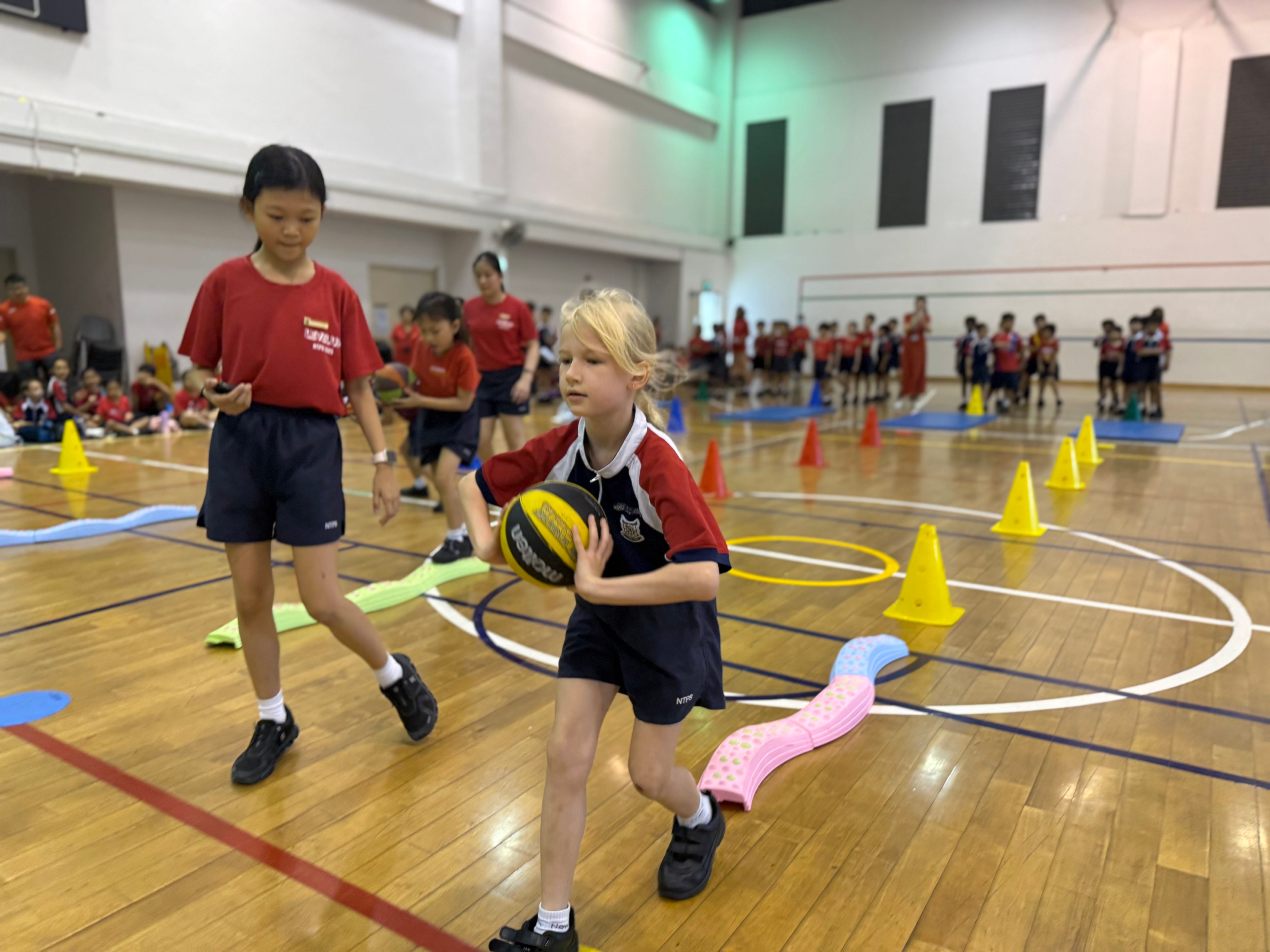 A young girl holds a basketball on an indoor gym court with colorful cones and hoops while other children watch.