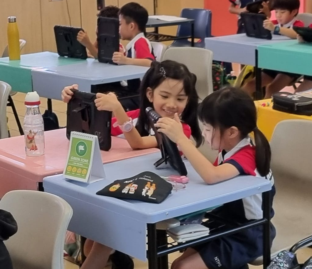 Two young girls in school uniforms share a tablet at a desk, smiling, with more students using tablets behind them.