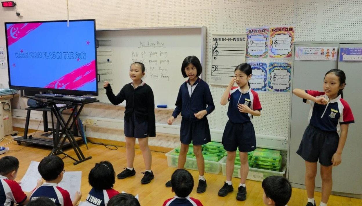 Four students in school uniforms perform in a music classroom while classmates watch, with a screen showing "Wave Your Flag in the Sun."