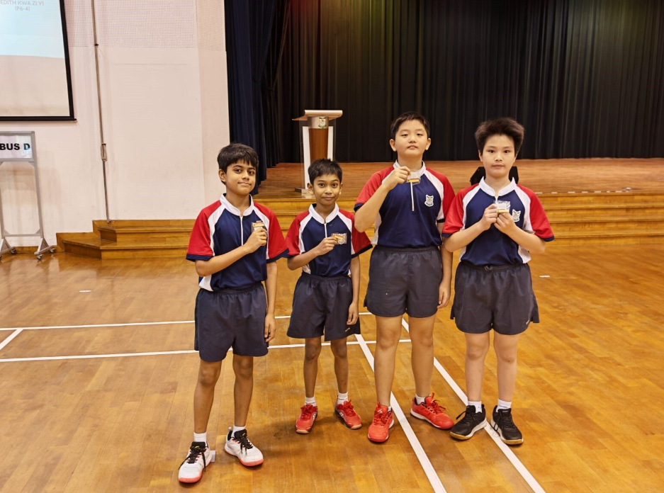 Four children in matching blue and red sports uniforms stand on a gymnasium floor, each holding a medal.
