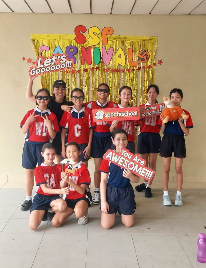 Group of students in red sports uniforms posing in front of a gold tinsel SSP Carnivals banner holding signs.