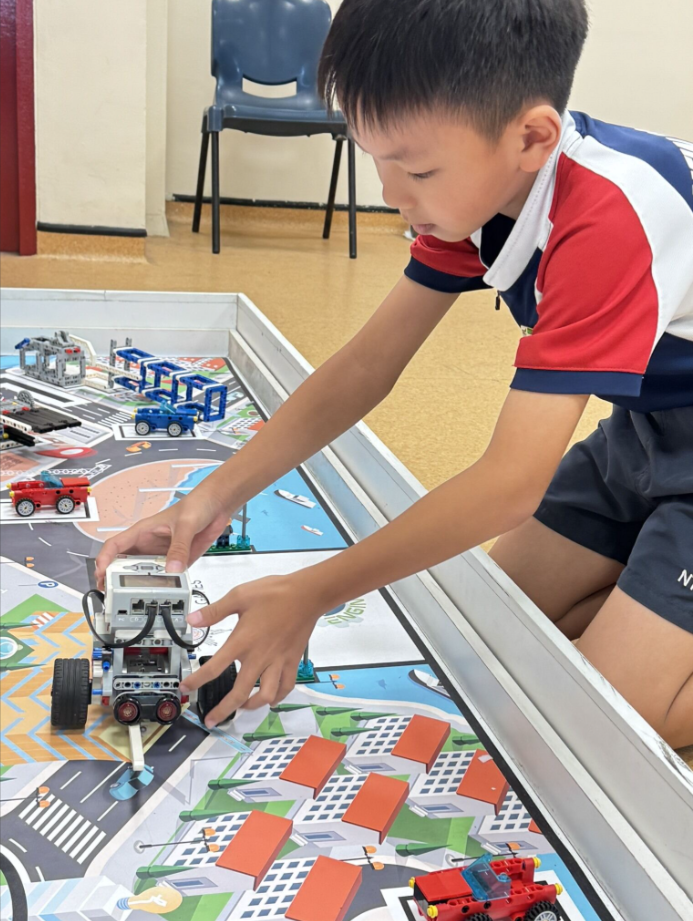 A young boy in a red and white shirt places a small robot on a colorful city-themed LEGO competition mat.
