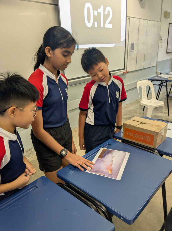 Three school children in uniforms examine a photo on a desk during a timed classroom activity showing 0:12.