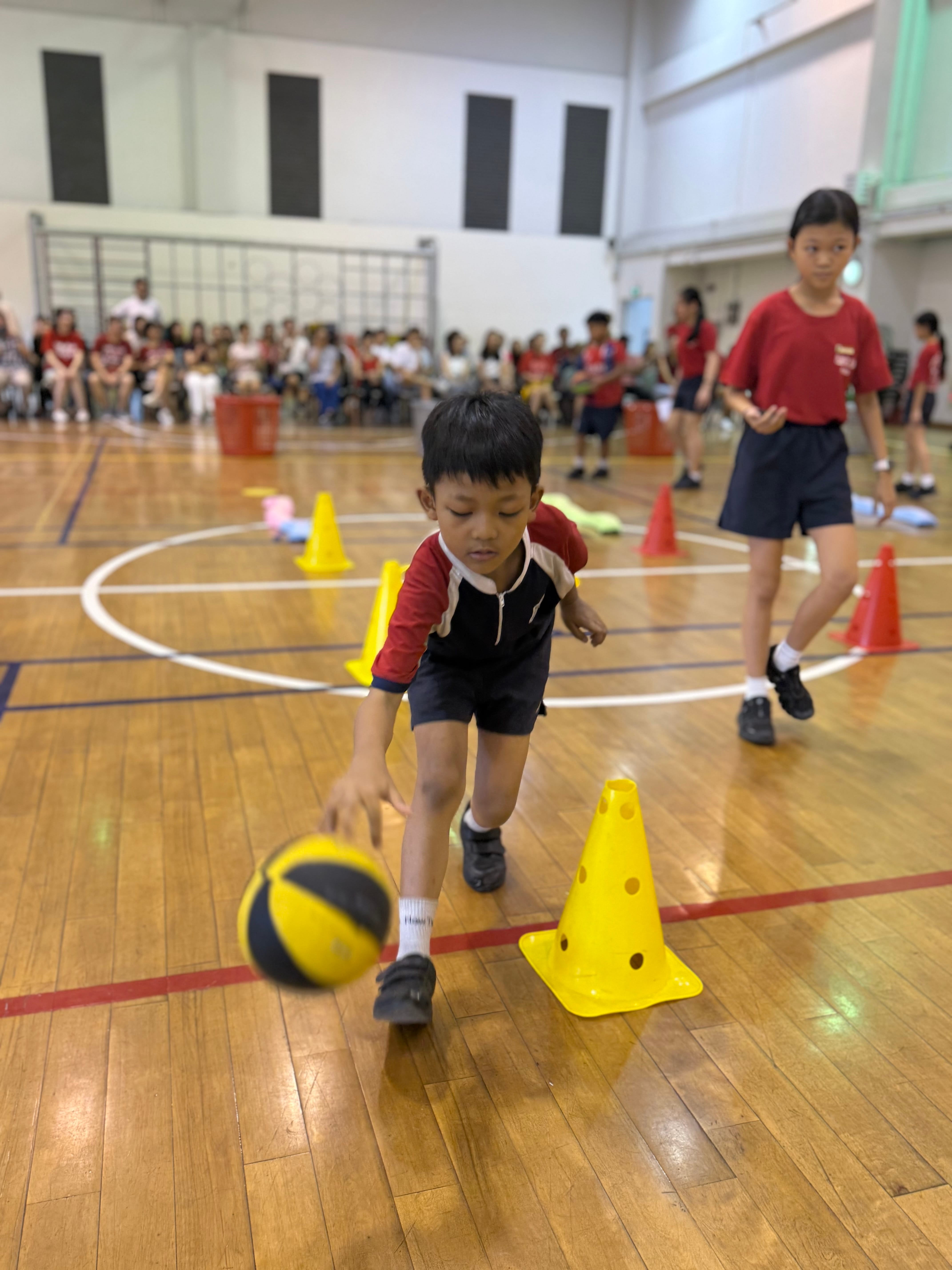 A young boy dribbles a ball around yellow cones on a gym floor while other children watch from the background.
