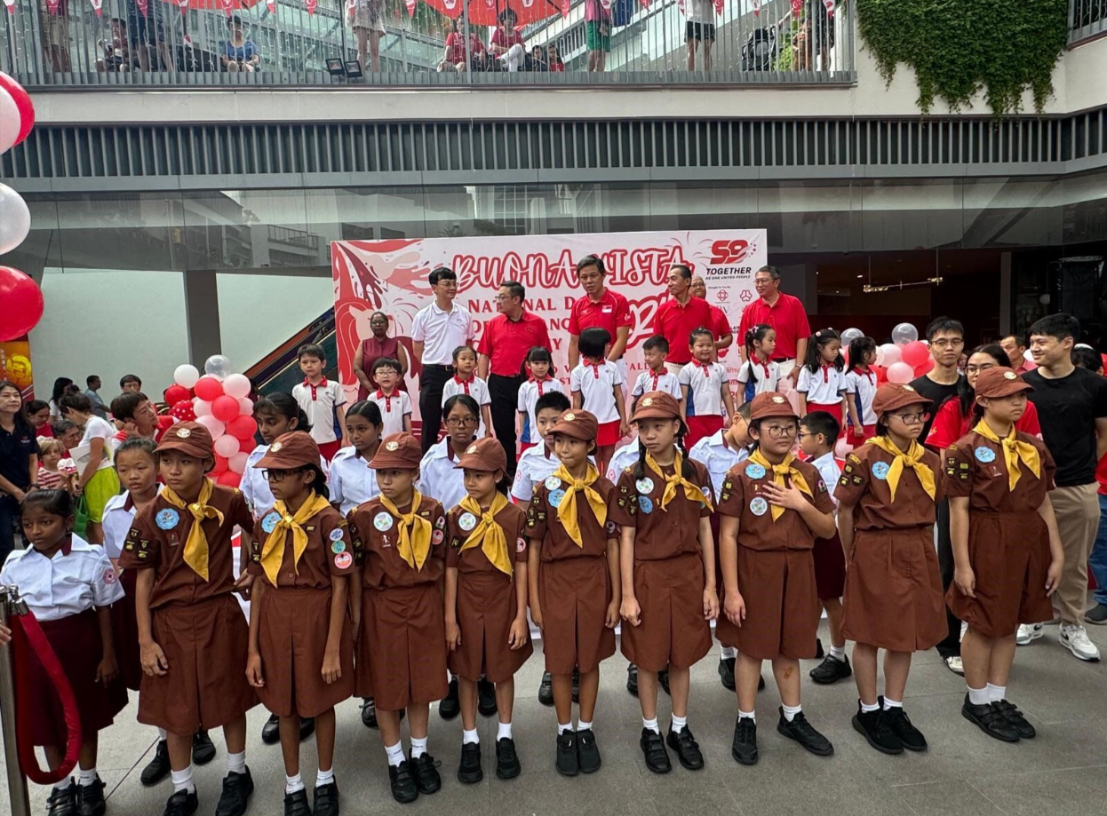Children in brown scout uniforms with yellow neckerchiefs stand at a Buona Vista National Day celebration event with red balloons.