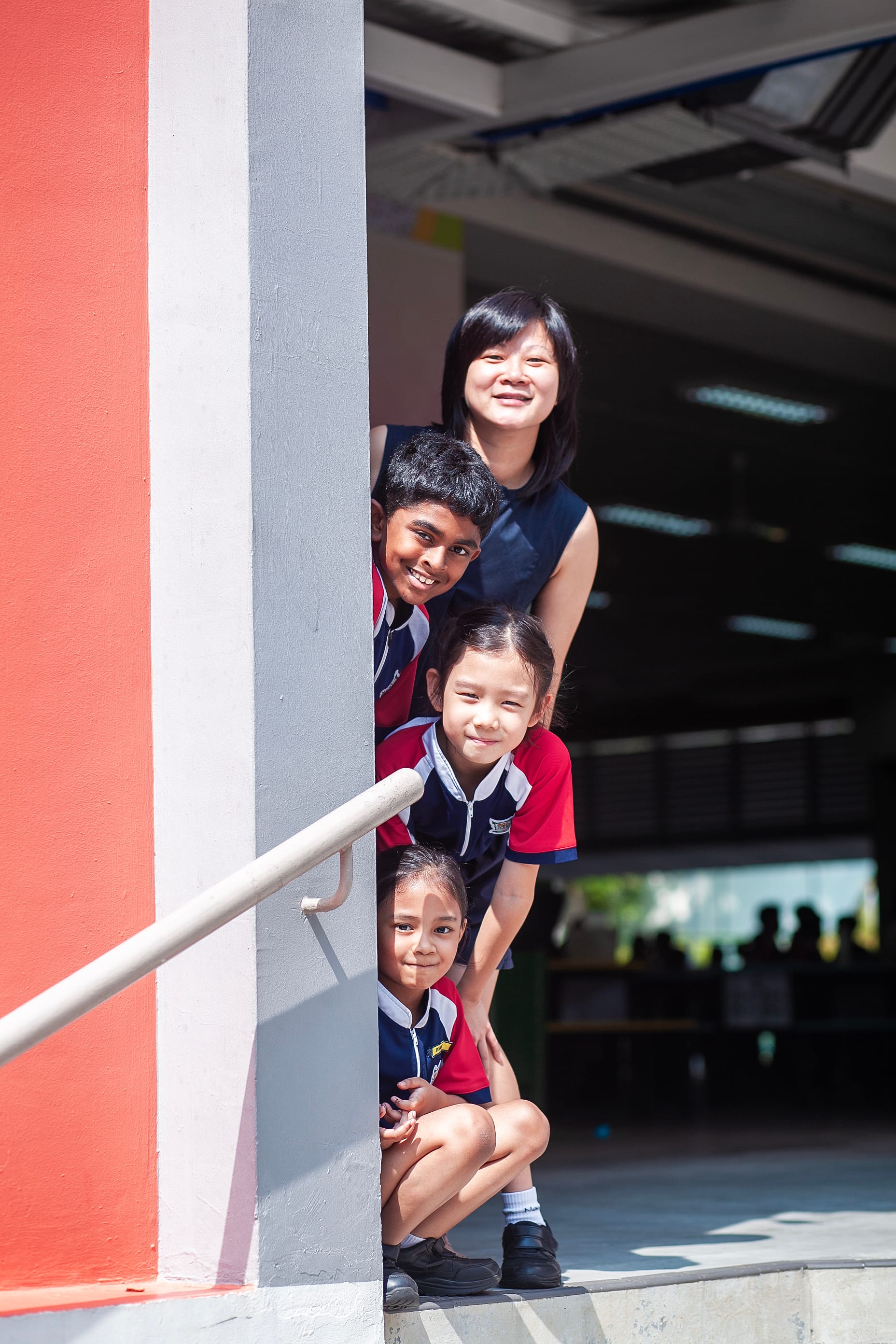 A smiling woman and three children in school uniforms peek playfully around a pillar at a school building.