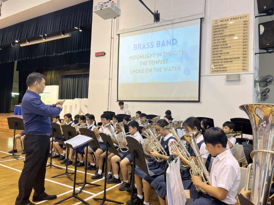 A conductor leads a school brass band of young students playing trumpets in a hall with a projected slide.