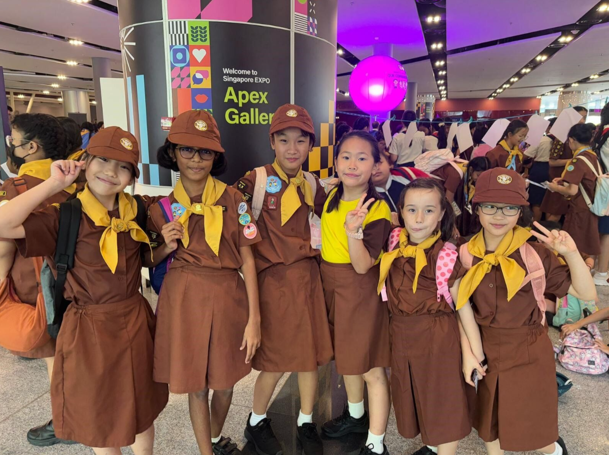 Six children in brown scout uniforms with yellow scarves pose at the Singapore EXPO Apex Gallery entrance.