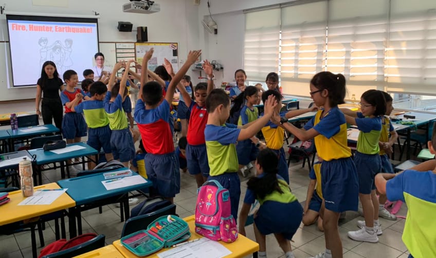 Children in blue uniforms play an activity game in a classroom with a "Fire, Hunter, Earthquake!" slideshow on screen.
