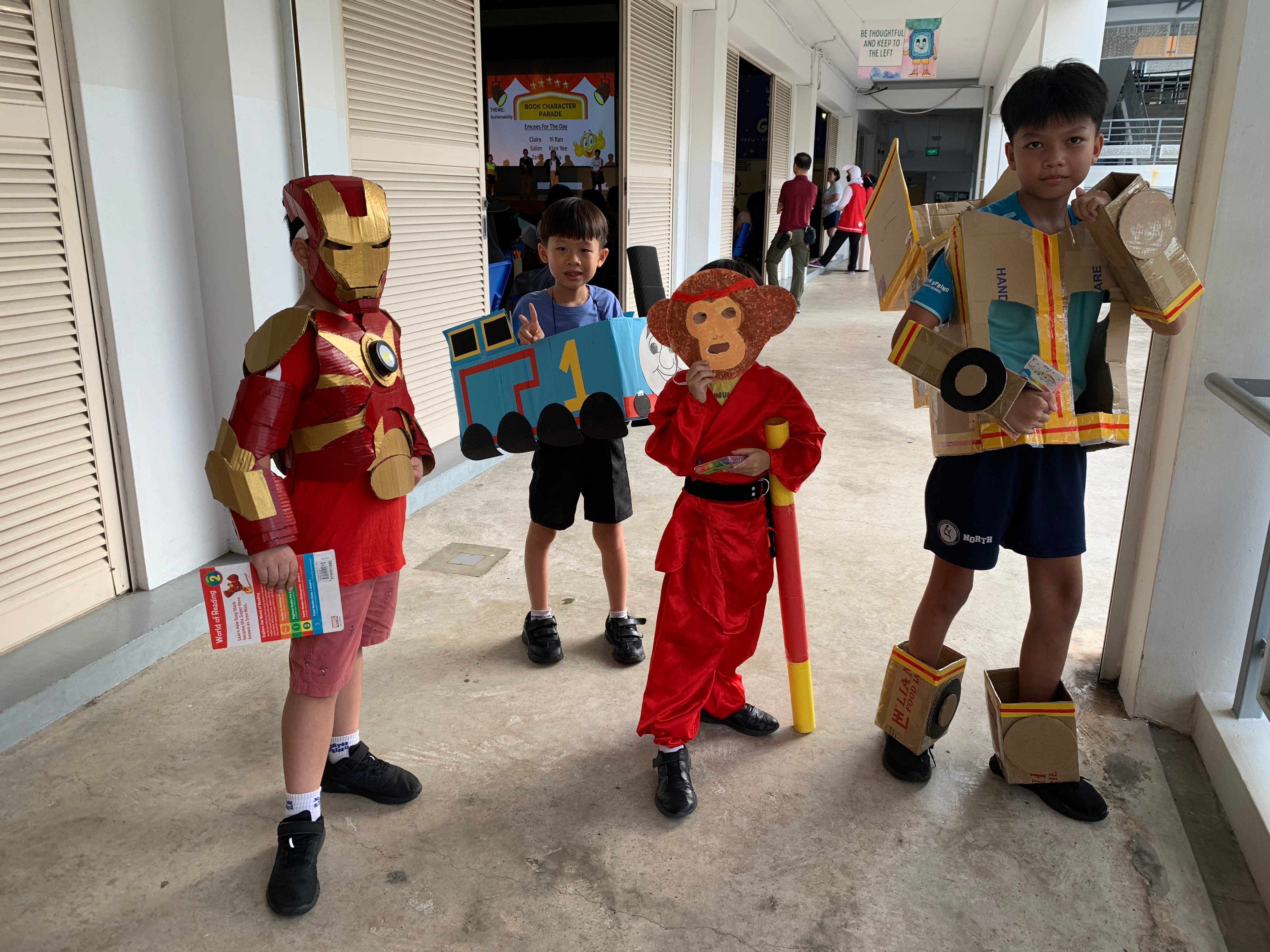 Four children in book character costumes including Iron Man, monkey king, and cardboard robot at a school parade.