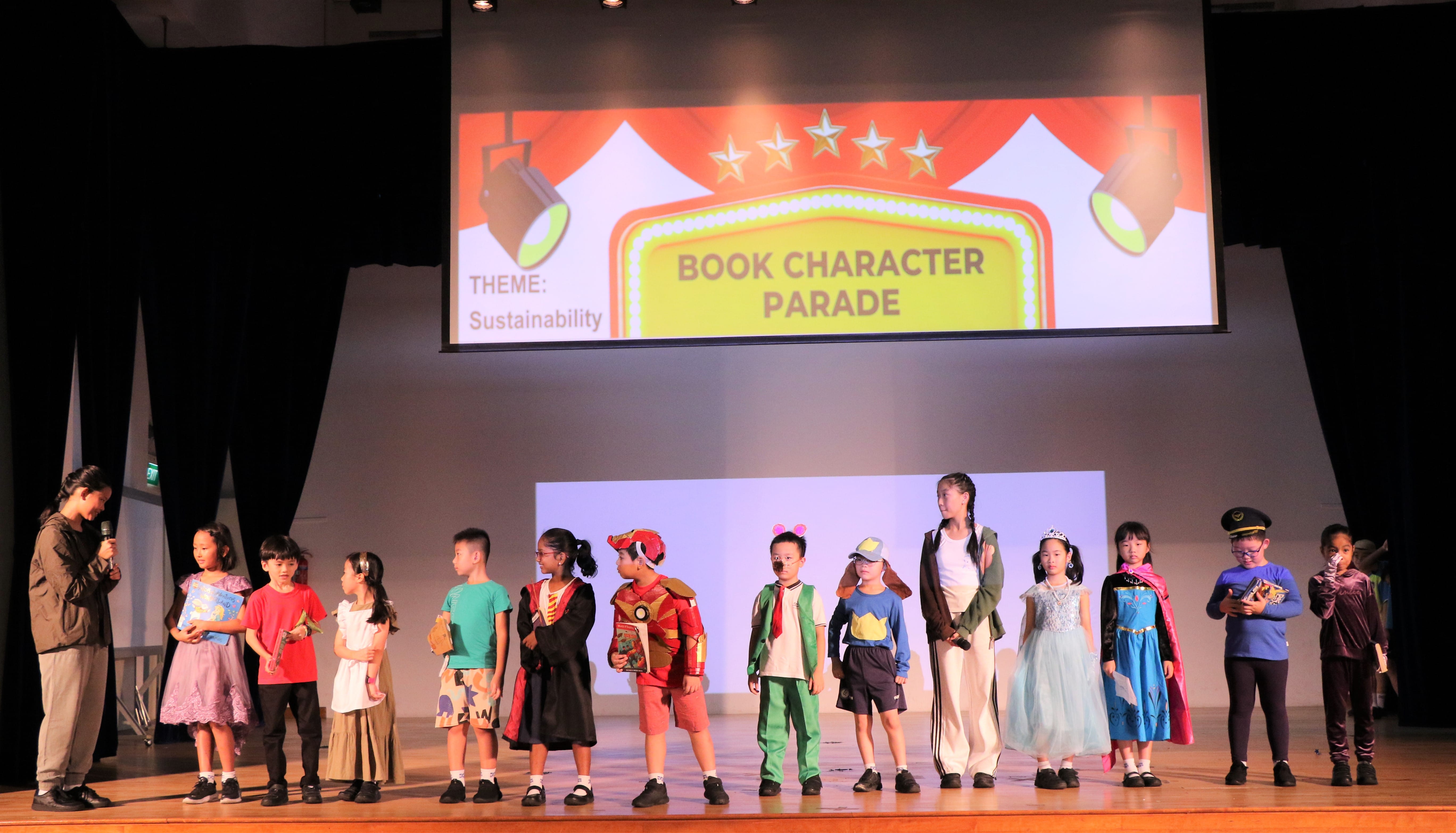 Children in costumes stand on stage at a Book Character Parade event with a sustainability theme projected behind them.