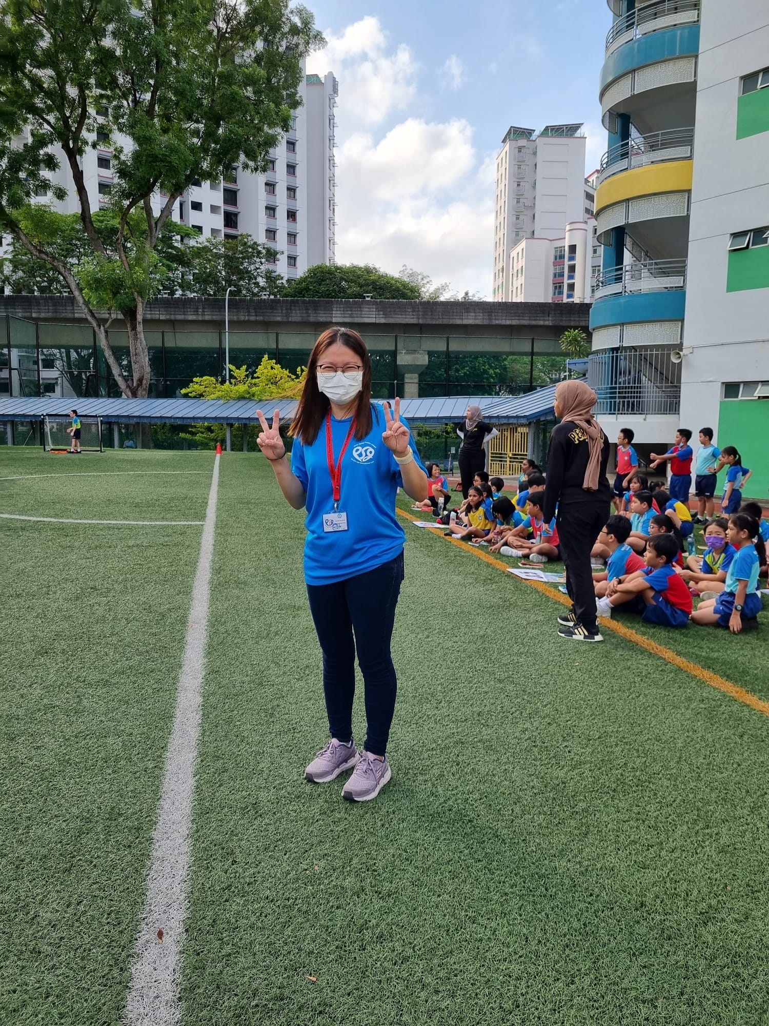 Woman in blue shirt and face mask making peace signs on a school field with children seated nearby.