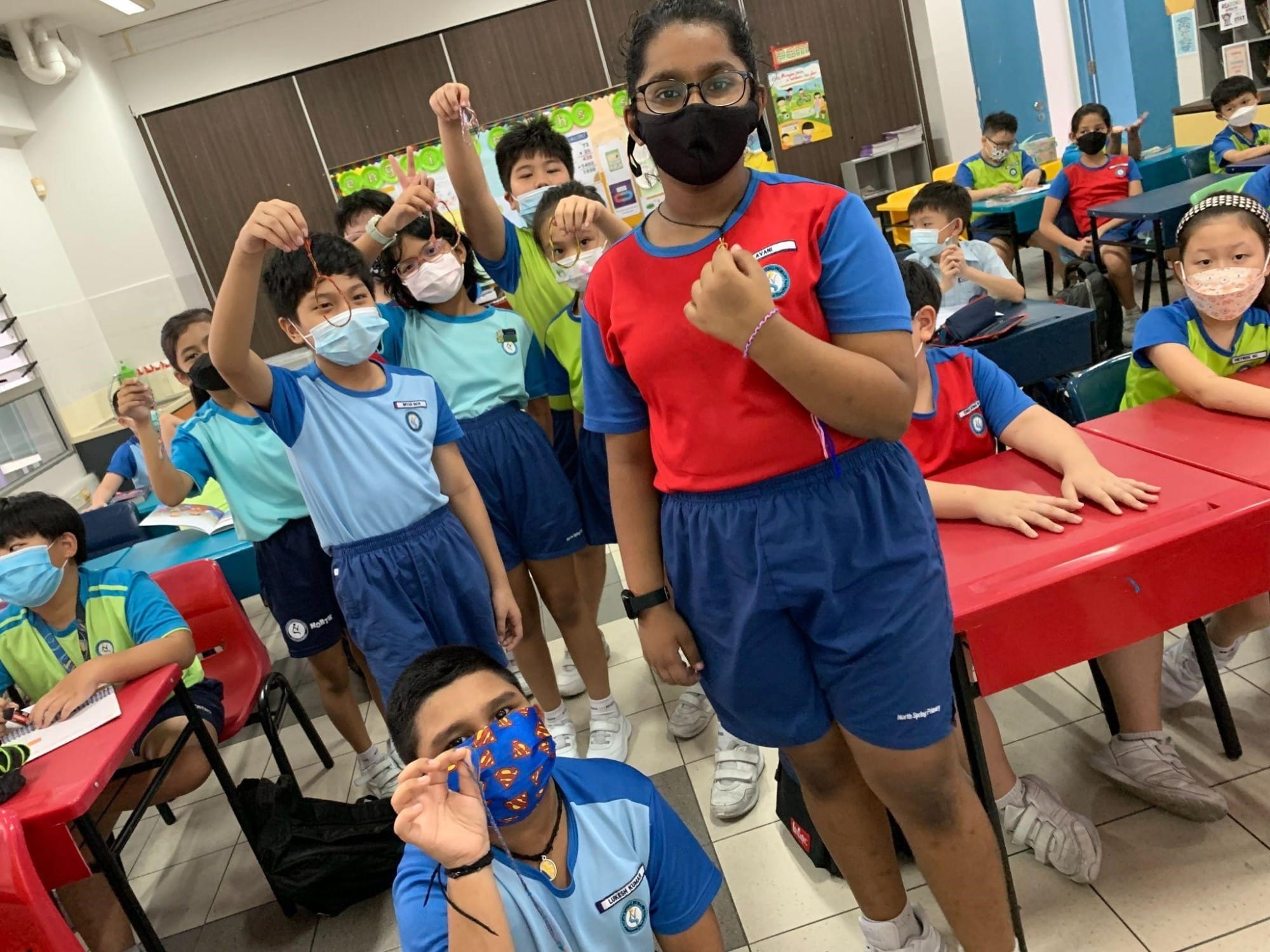 Children in school uniforms and face masks stand and sit in a classroom, some holding up small craft items.