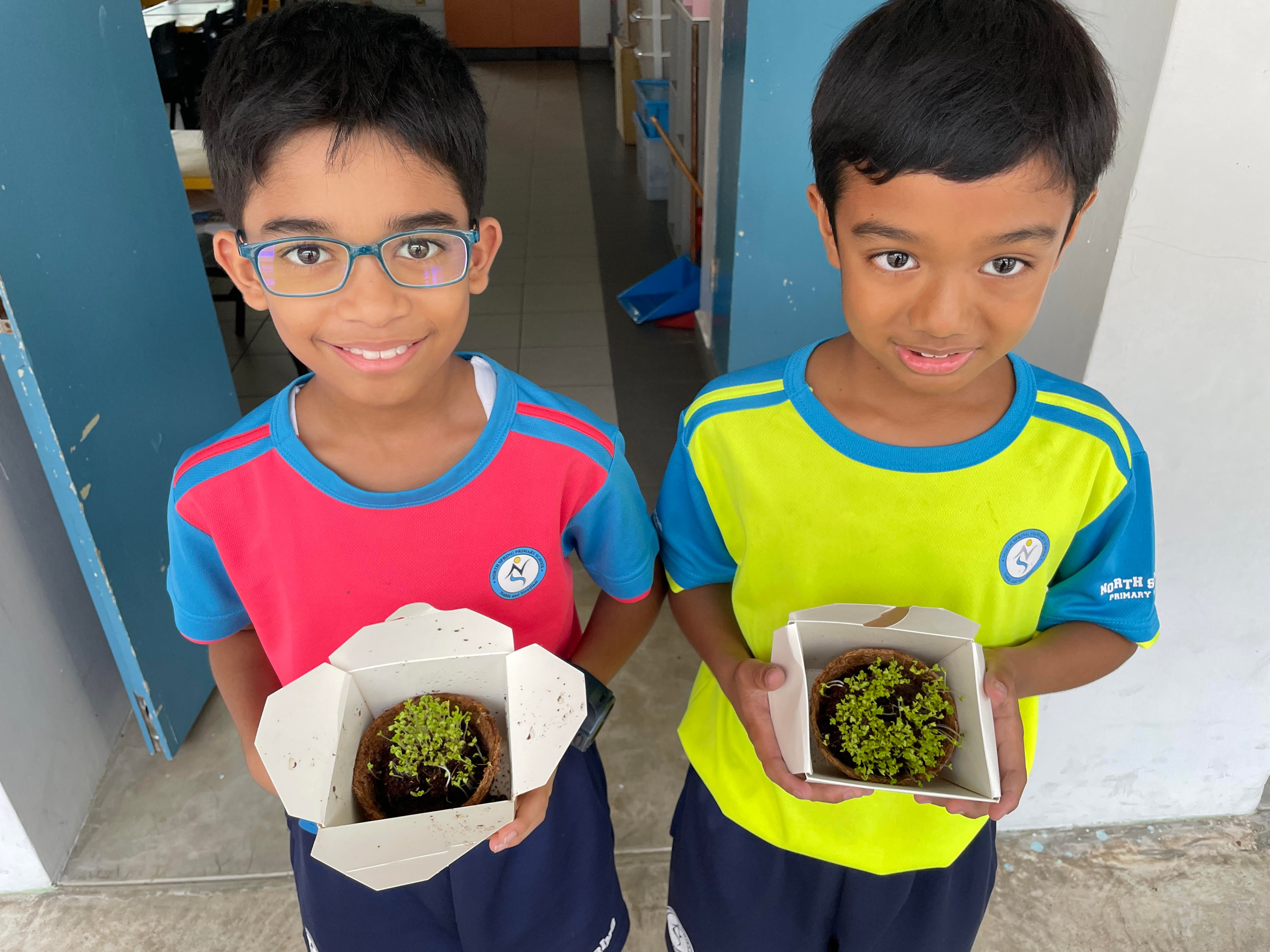 Two young boys in school uniforms each hold a small cardboard box containing a sprouting seedling in a coir pot.