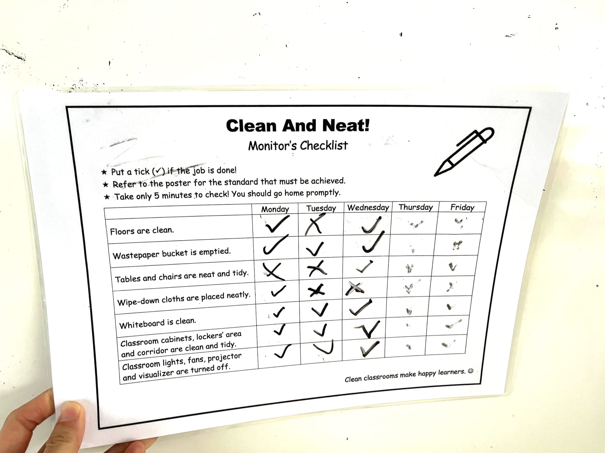 A hand holds a classroom monitor checklist titled "Clean And Neat!" with daily tick marks for cleaning tasks.