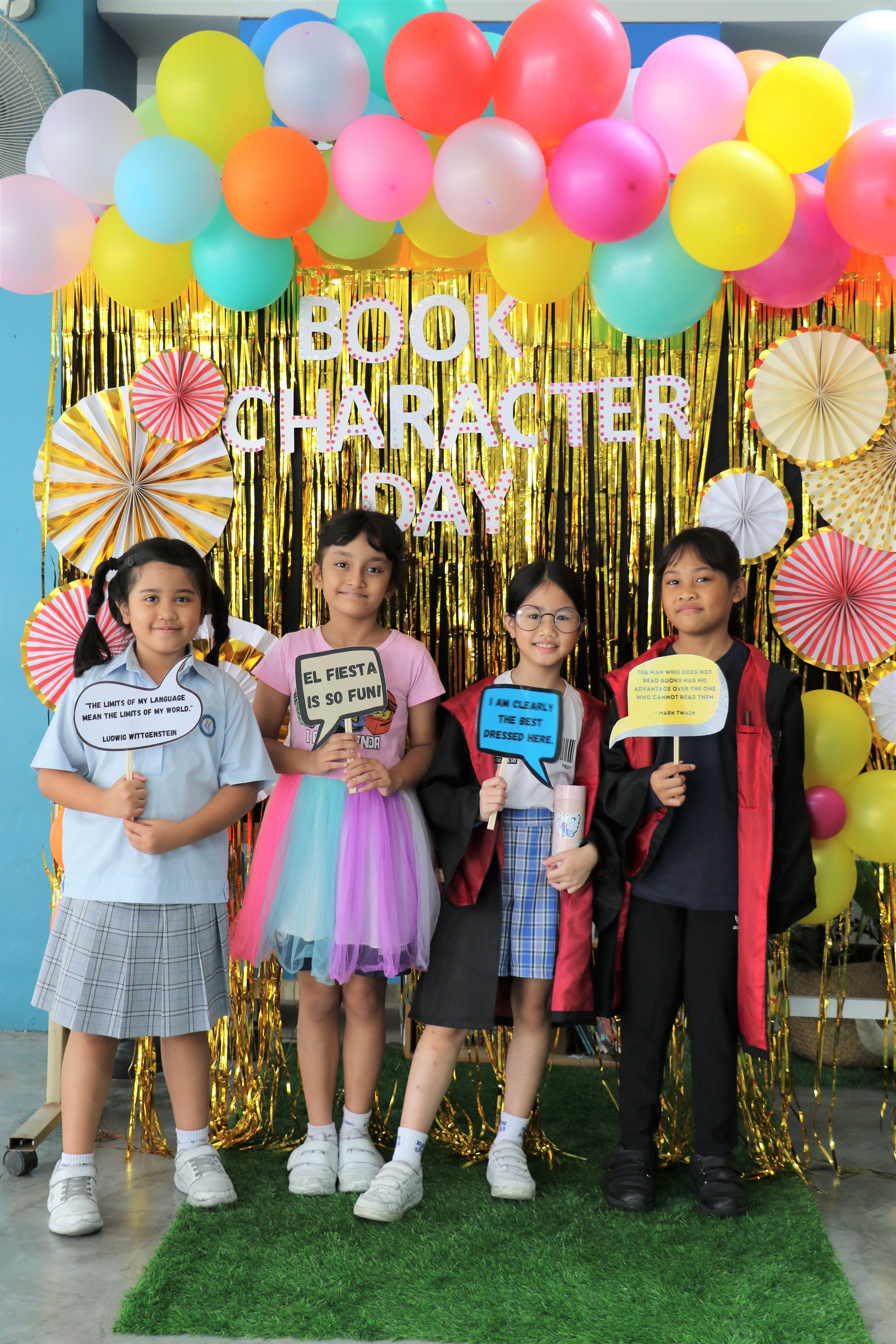 Four children hold quote signs in front of a colorful balloon arch and gold fringe backdrop reading Book Character Day.