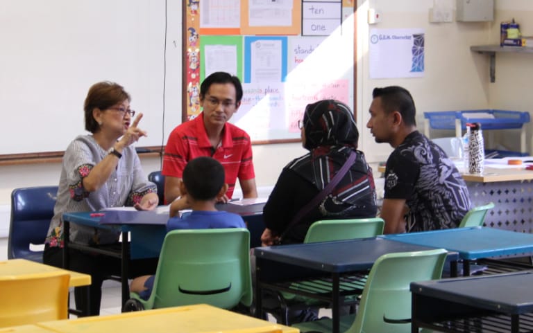 A teacher speaks with three adults and a child seated around desks in a classroom with colorful wall displays.