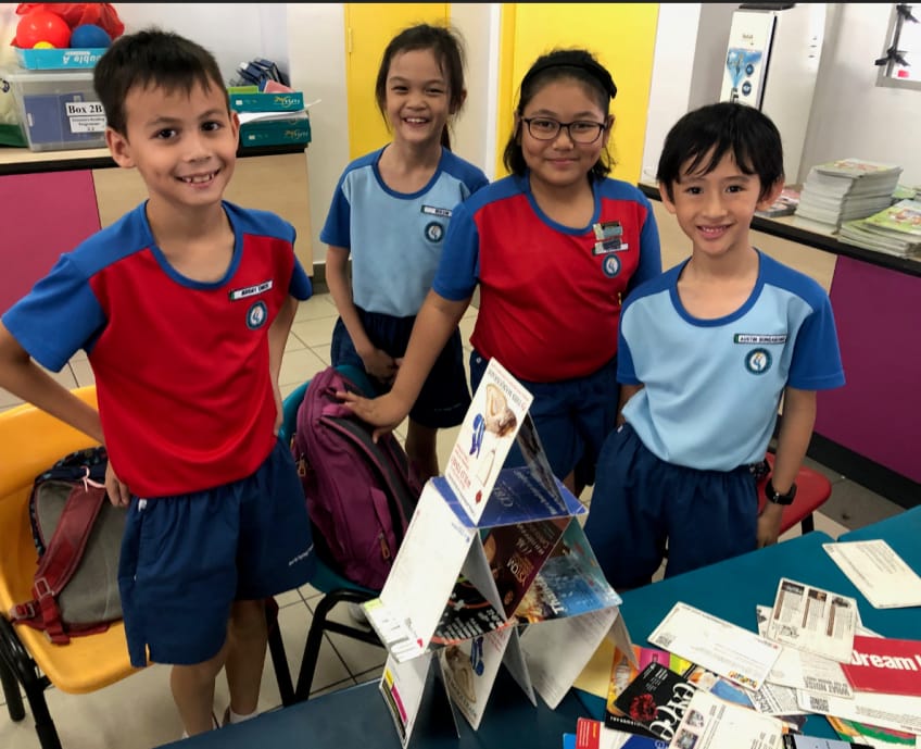 Four smiling children in blue and red school uniforms stand near a table with books and papers in a classroom.