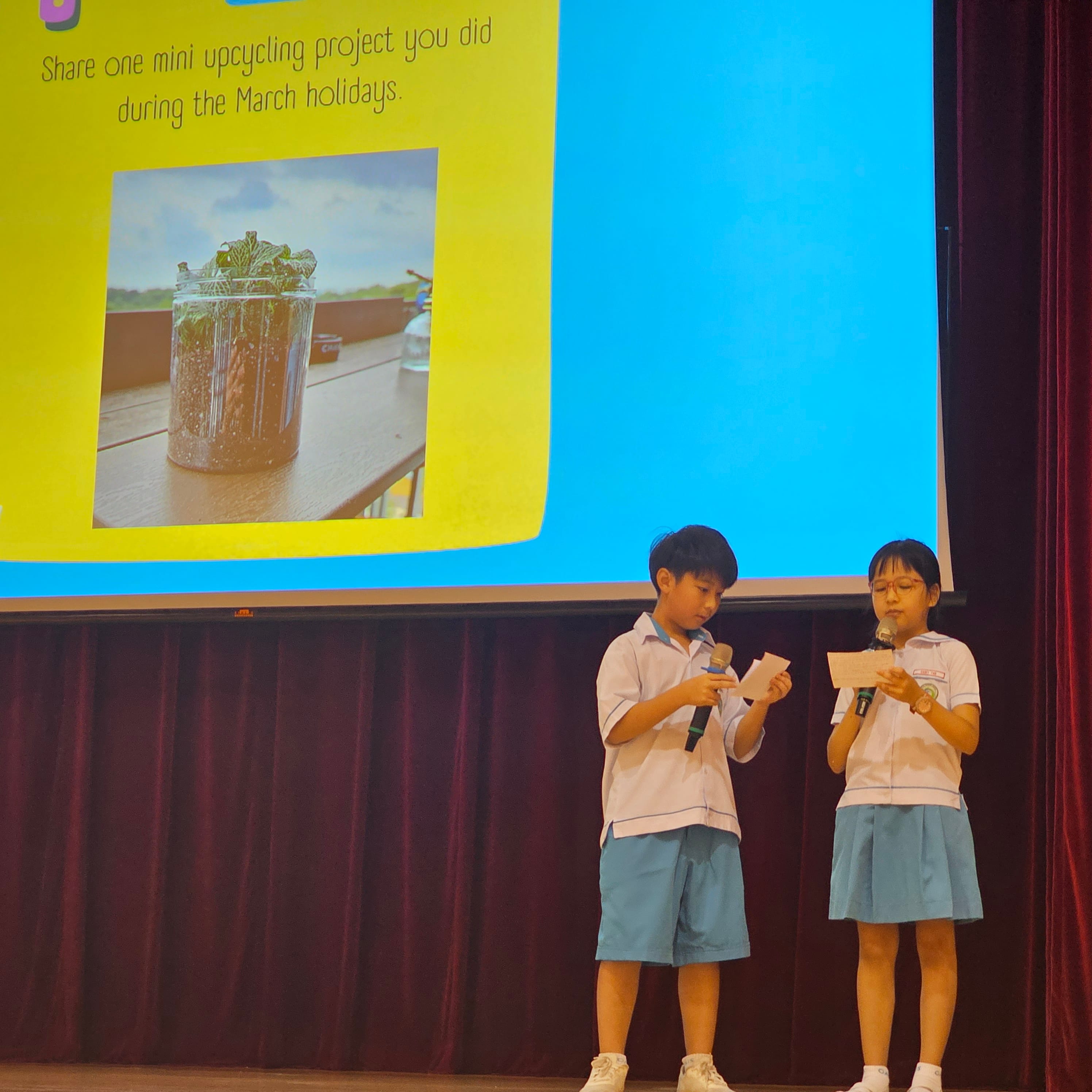 Two uniformed schoolchildren hold microphones on stage in front of a projected upcycling project slide.