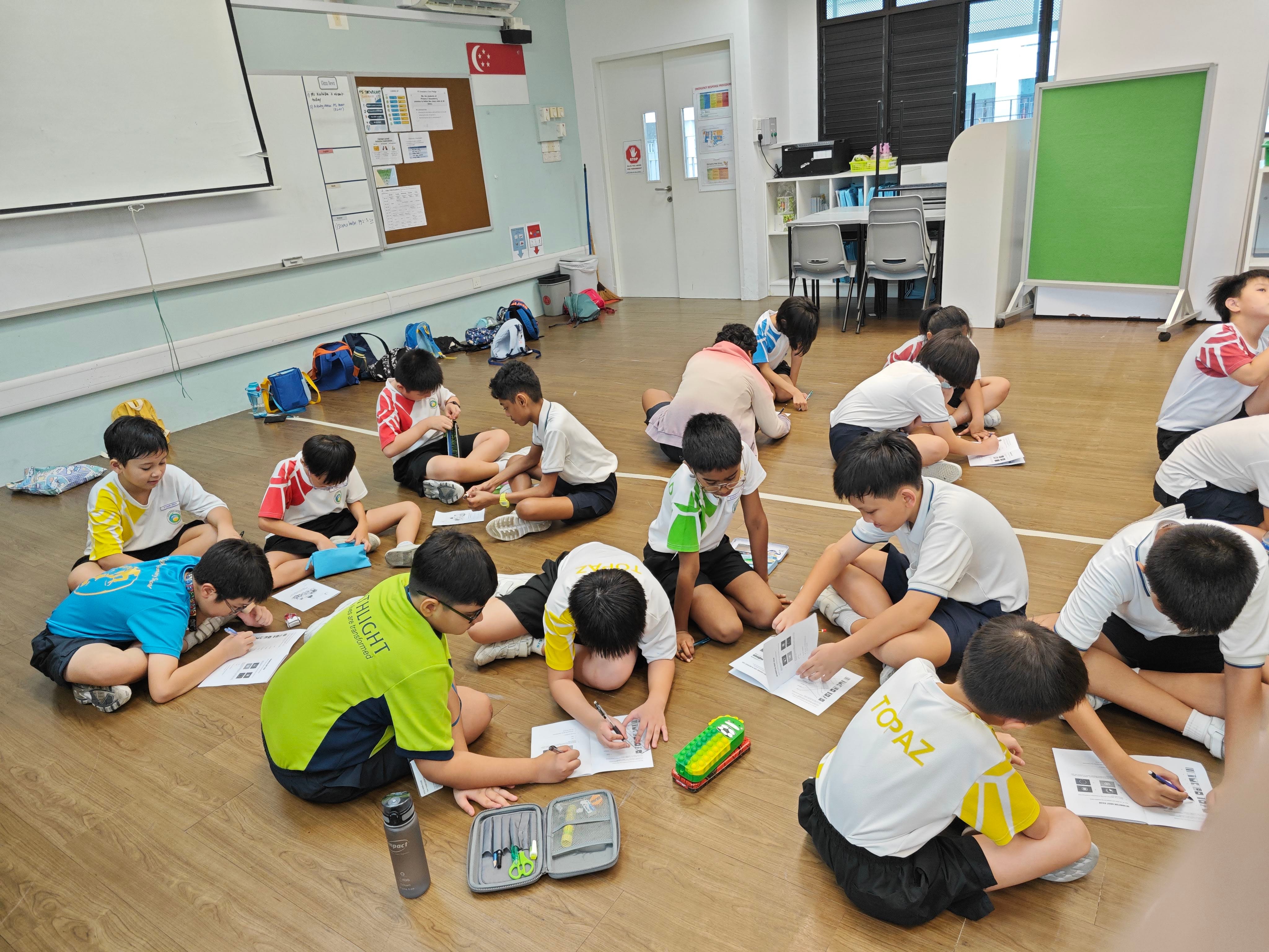 Students in school uniforms sit on a classroom floor writing on papers during a group activity.