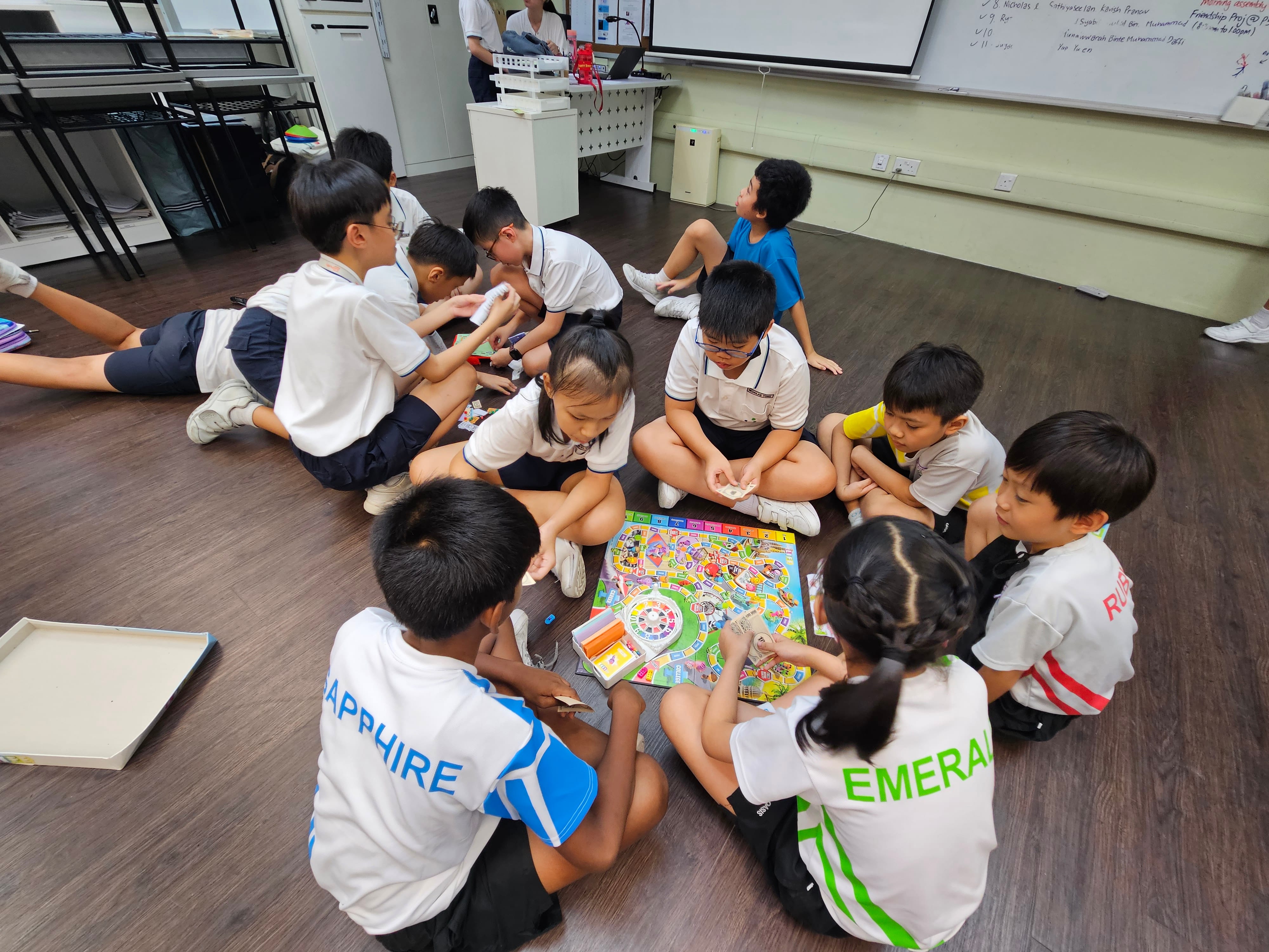 Children in school uniforms sit on a classroom floor playing a colorful board game together.