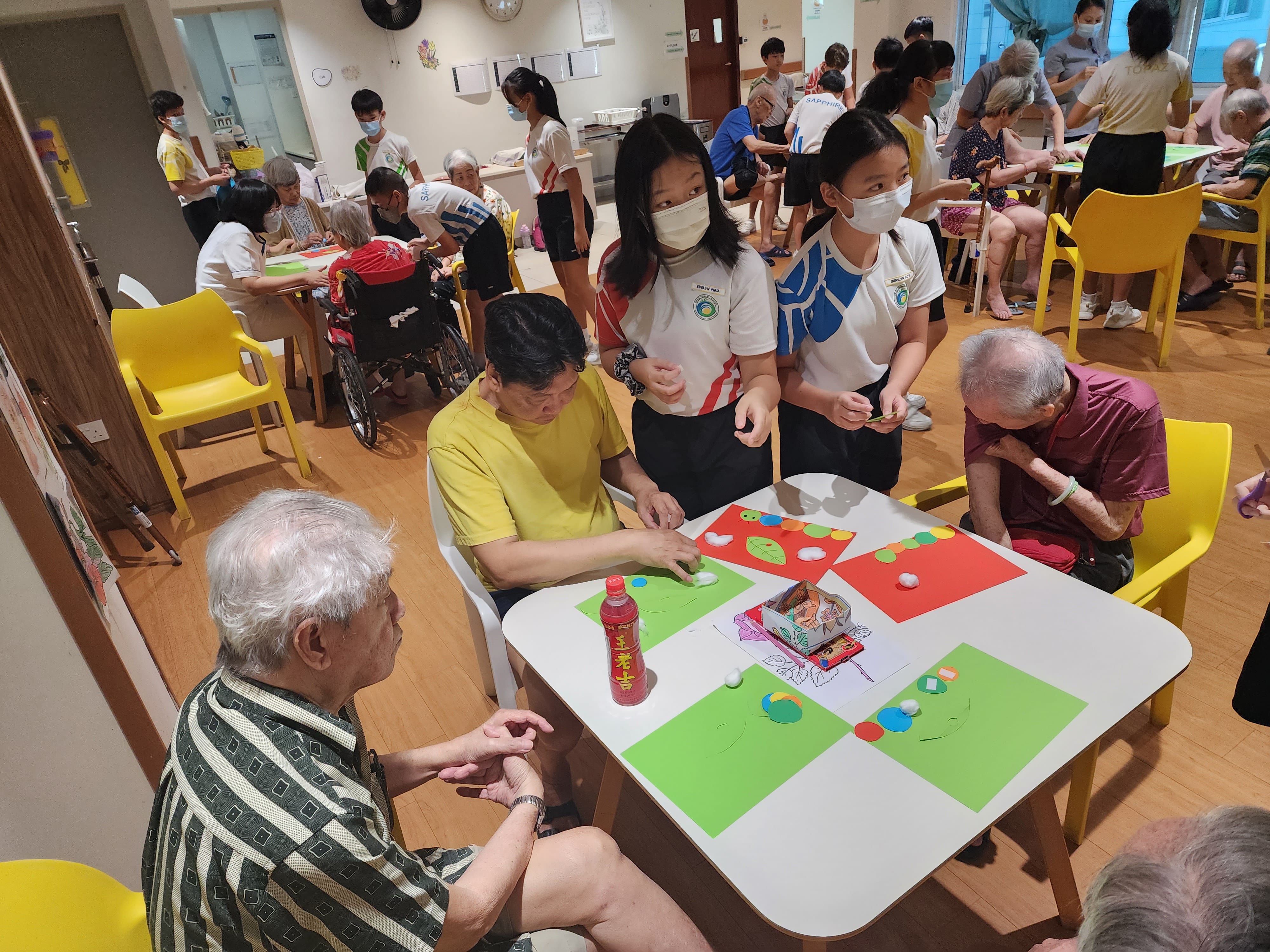 Young masked students assist elderly people with a colorful paper craft activity in a busy care center room.