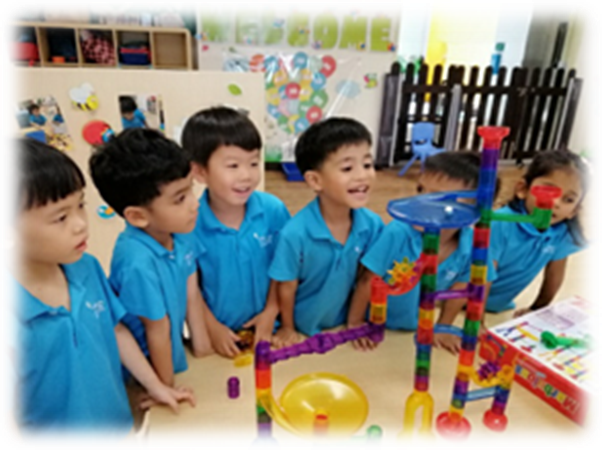 Six young children in blue uniforms stand around a table with a colorful building block tower in a classroom.
