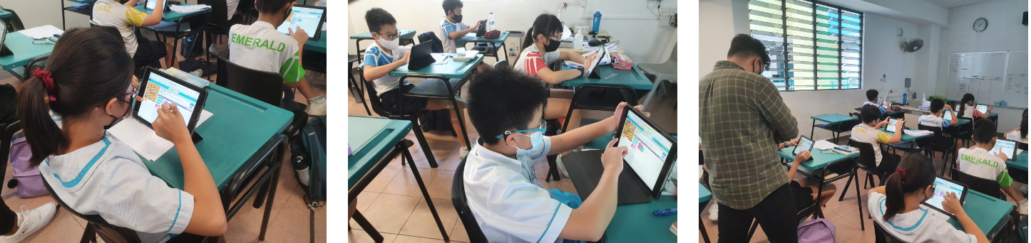 Students in school uniforms and masks use tablets at green desks in a classroom, with a teacher assisting.