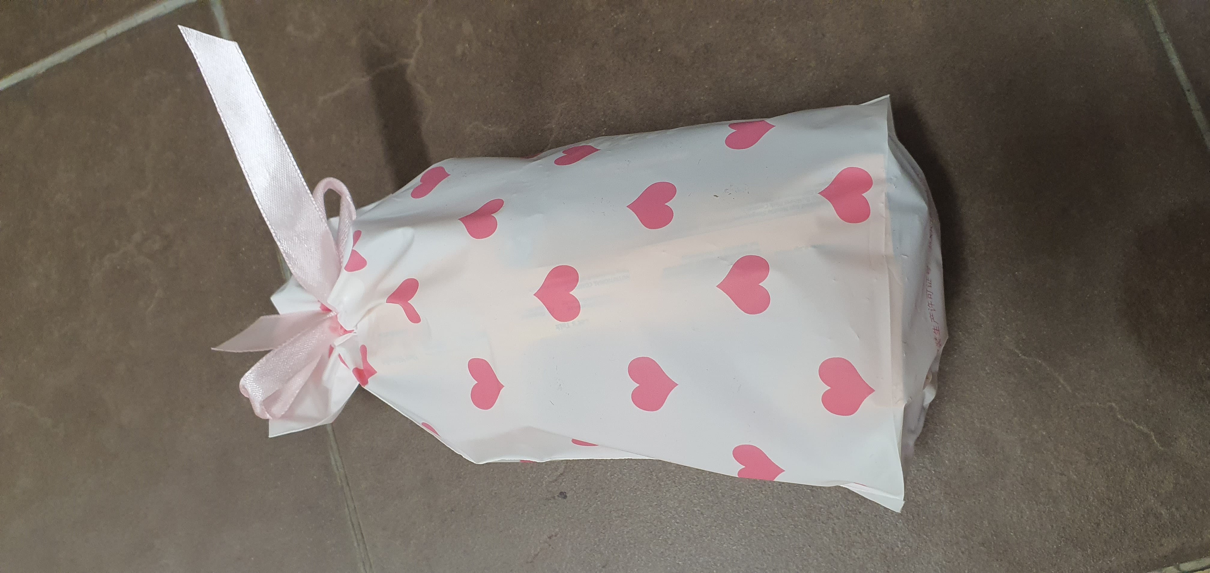 White gift bag covered in pink hearts, tied at the top with a pink satin ribbon, resting on a gray tiled floor.